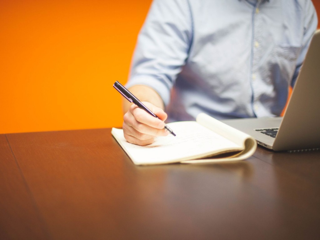 Une main prend des notes sur un carnet, à côté d'un ordinateur portable ouvert sur un bureau en bois.