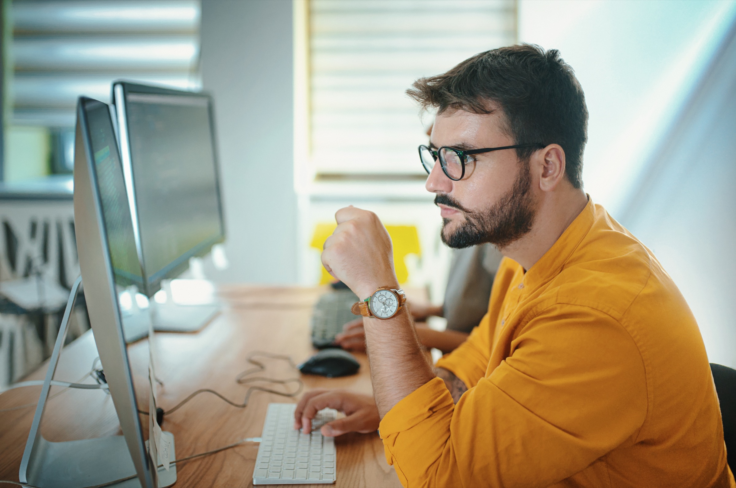 Une personne installée à son poste de travail, devant deux écrans, dans un bureau éclairé par la fenêtre.